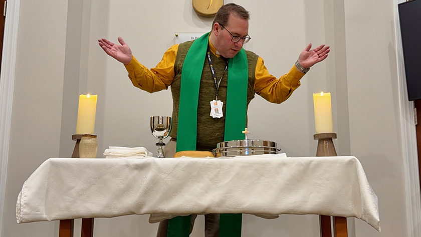 A person wearing a yellow shirt and green stole stands behind a table covered with a white cloth, arms extended outward. On the table are two lit candles on wooden holders, a silver chalice, folded white cloths, a loaf of bread, and a stack of silver communion trays. Behind the person is a wall with a large wooden cross featuring decorative carvings and the letters "SP".