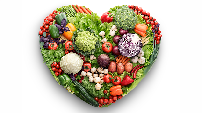 A photograph of various fresh vegetables arranged in a heart shape on a white background, symbolizing healthy eating and nutrition. The assortment includes leafy greens, tomatoes, carrots, broccoli, mushrooms, and peppers, showcasing vibrant colors and diverse textures.