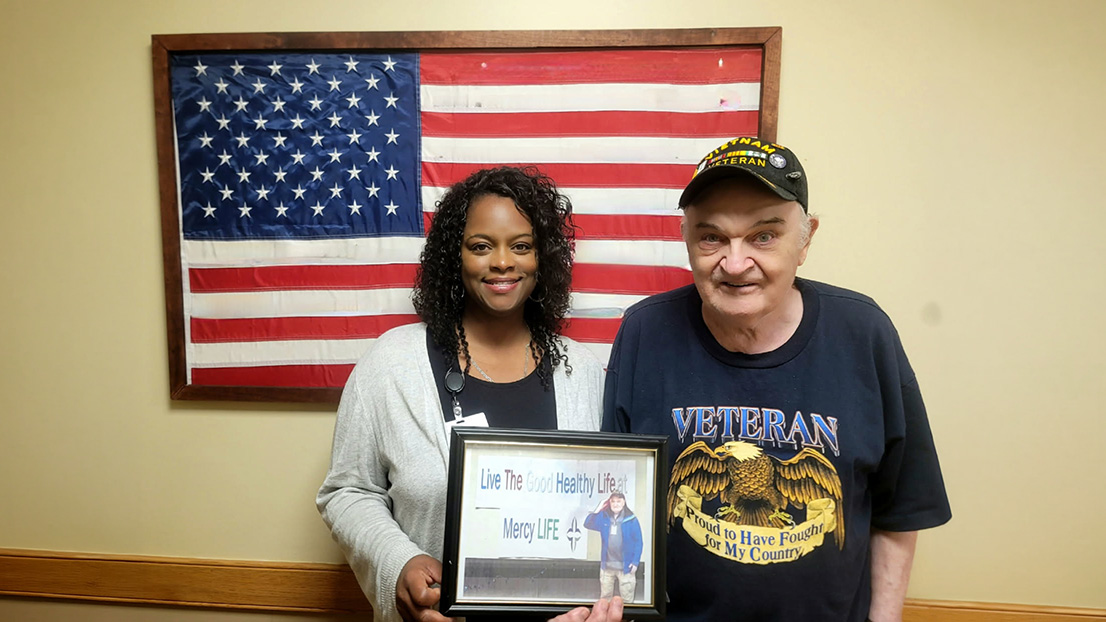 Participant and staff member displaying a framed recognition photo in front of an American flag.